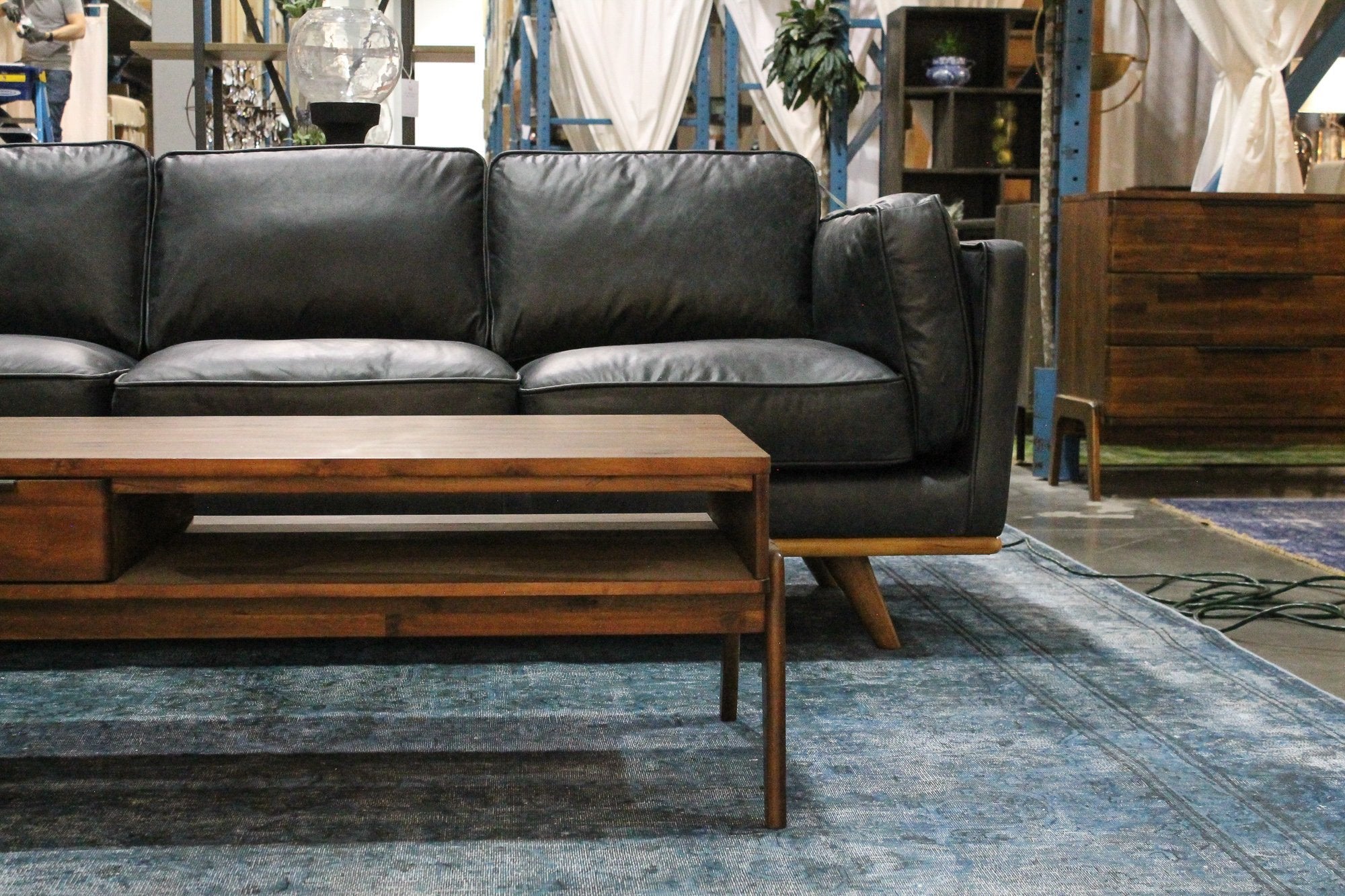 Remix Coffee Table placed in front of a black leather sofa in a showroom setting, showing the open lower shelf and warm brown wood tone